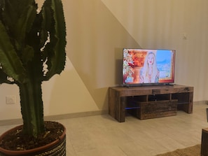 A living room with a television on a wooden stand, a large potted plant, and a tiled floor. - Maison nomade (3-star accommodation) (Feignies)