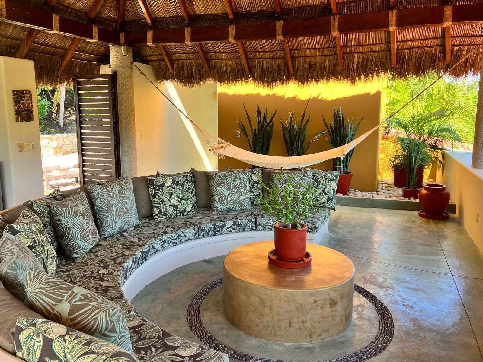 A curved sofa with patterned cushions, a round coffee table, a hammock, and potted plants in a sunlit room with a thatched roof.