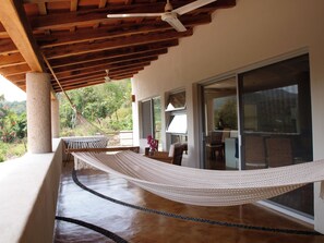 A hammock under a wooden ceiling, a glass door leading to a balcony, and a view of greenery outside.