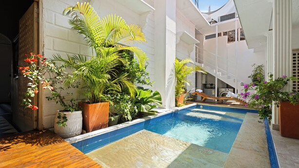 Una piscina con bordo in legno, circondata da piante in vaso e un edificio bianco con un balcone.