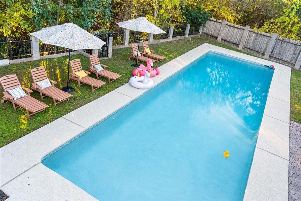 Ein rechteckiger Pool mit einer gefliesten Kante, umgeben von einer Rasenfläche mit Holzliegen und Sonnenschirmen.