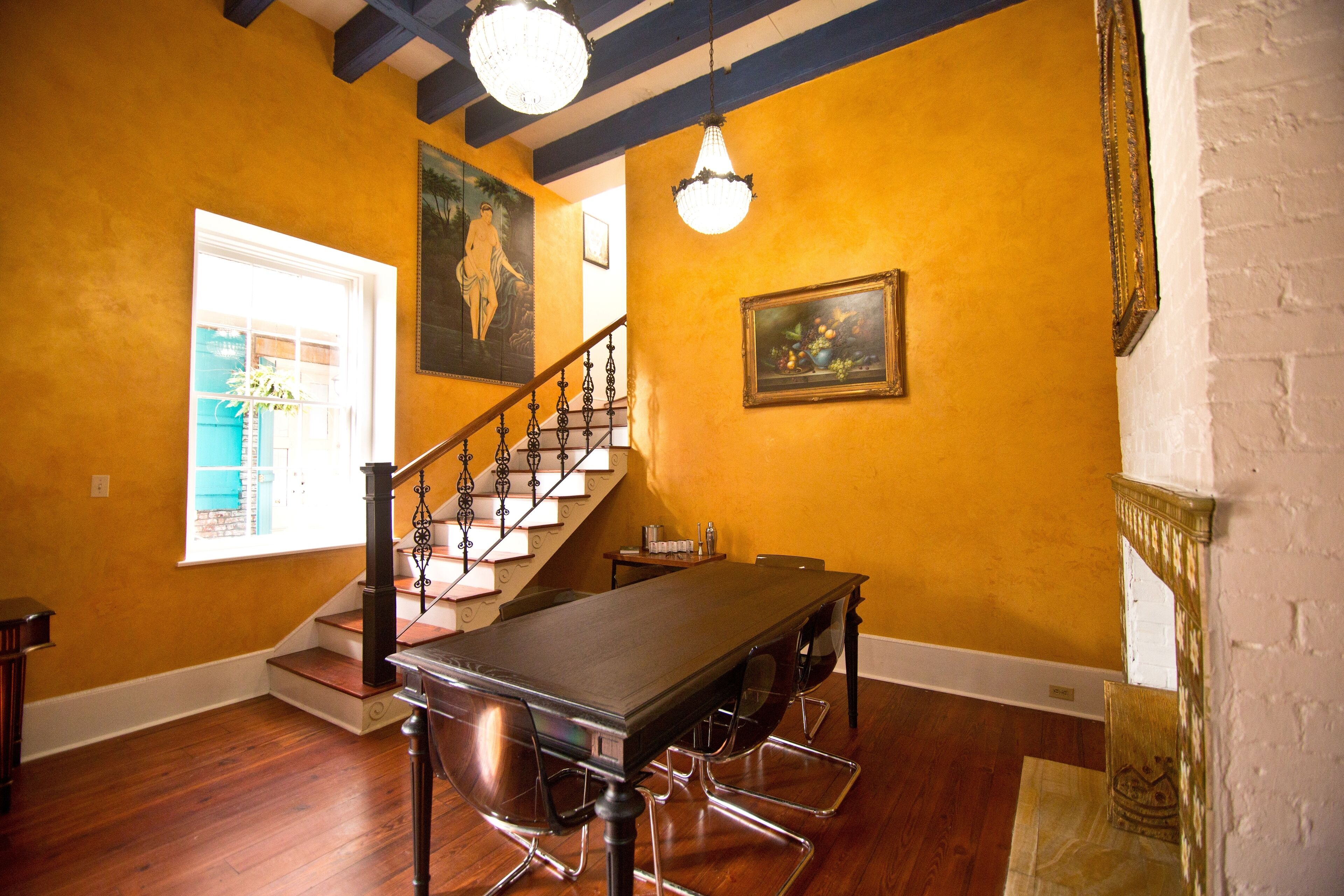 A spacious room with a wooden floor, a dining table with chairs, a staircase with a railing, and a chandelier.