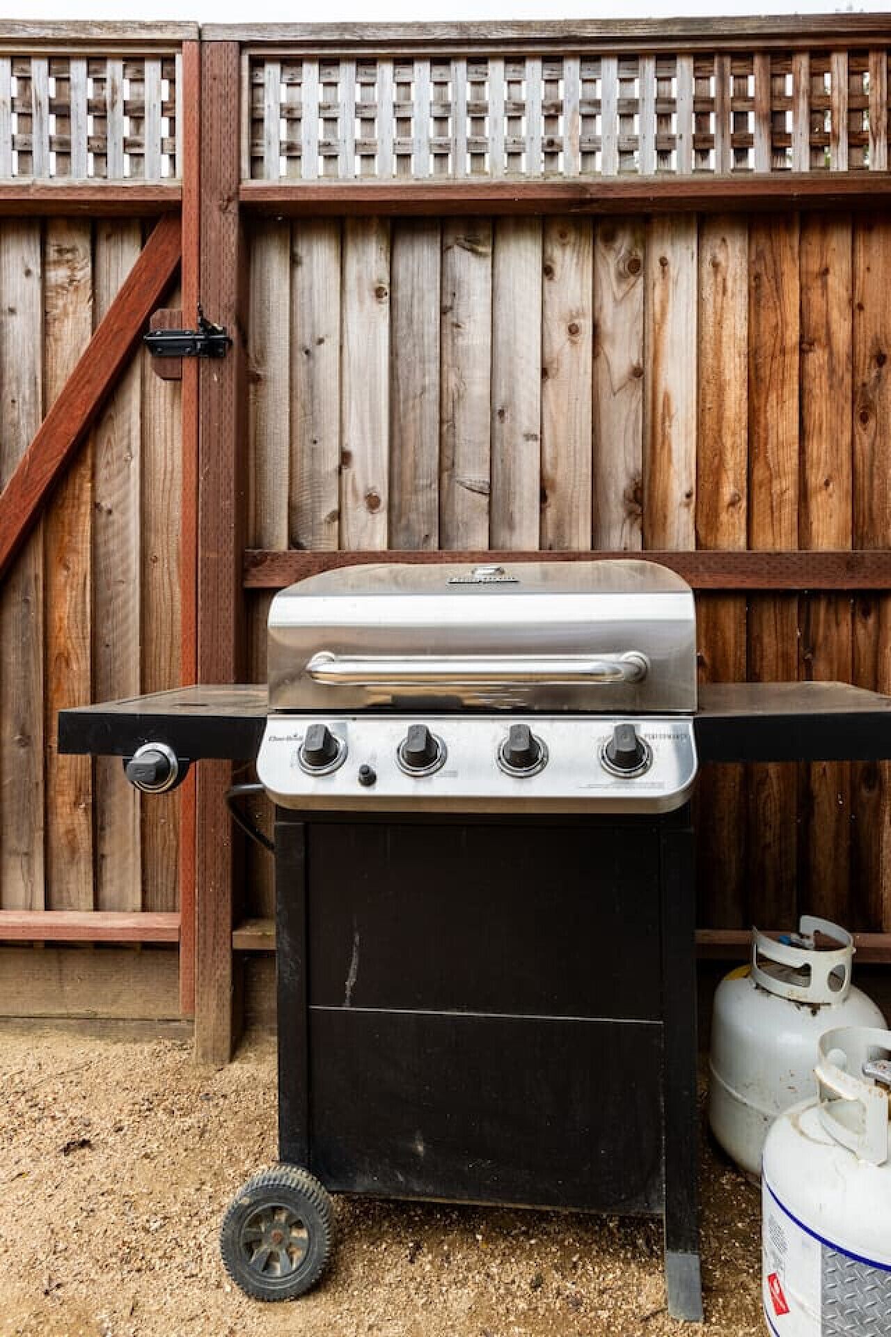 A portable gas grill with four burners, a side burner, and a storage compartment, set against a wooden fence.