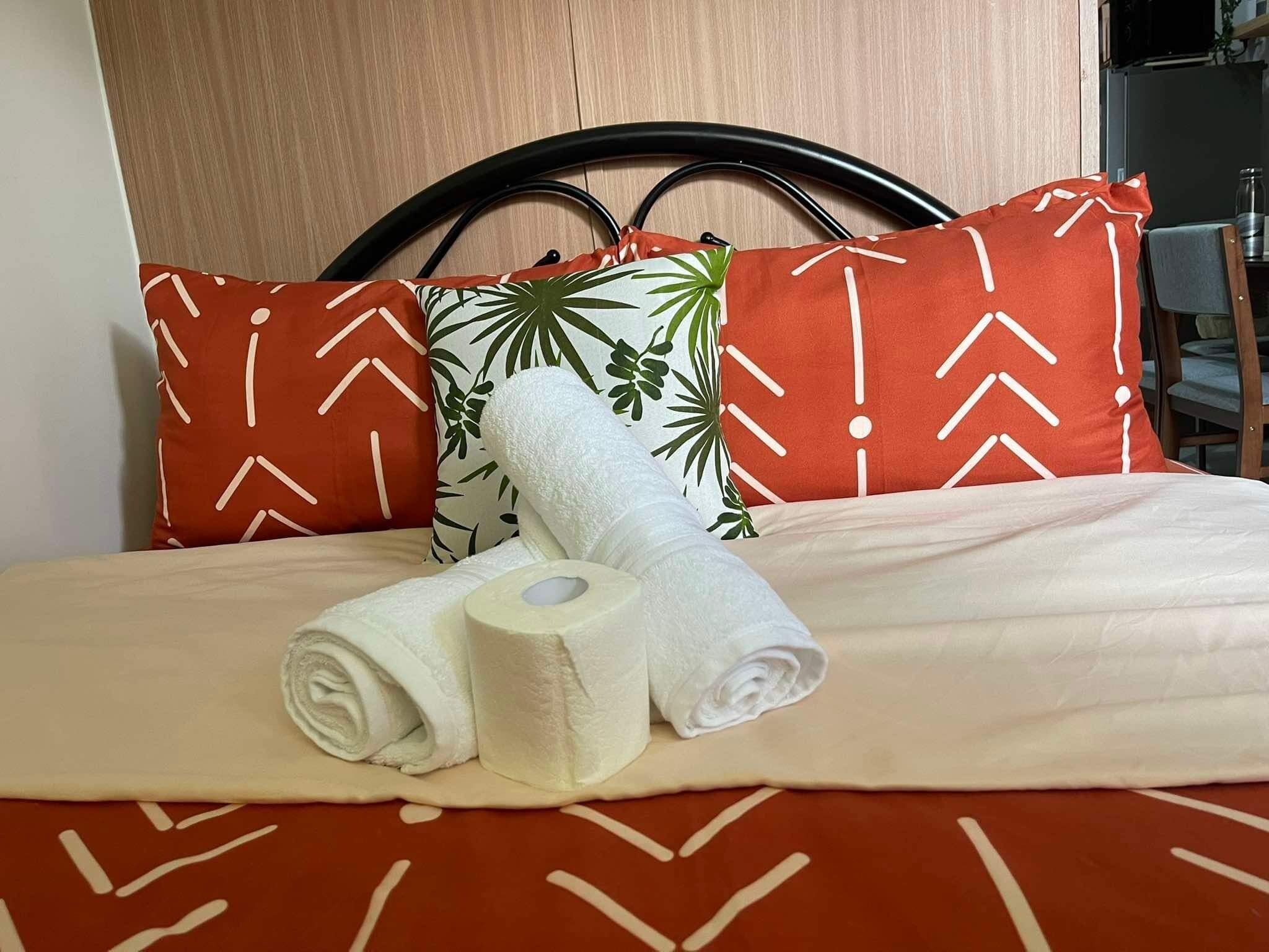 Een net opgemaakt bed met een beddekleed met dessin, twee opgerolde handdoeken en drie kussens met geometrische en bladmotieven.