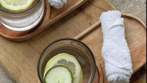 A wooden tray with two glasses of water, each with a slice of lemon and cucumber, and two rolled white towels.