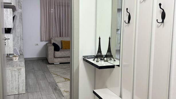 A modern interior with a hallway leading to a living area. There is a white wardrobe with black hooks, a mirror, and a small shelf with Eiffel Tower models.