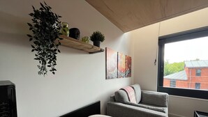 Un salon moderne avec un canapé gris, un fauteuil blanc, une œuvre d’art accrochée au mur, une étagère en bois ornée de plantes et une grande fenêtre donnant sur un bâtiment en briques.