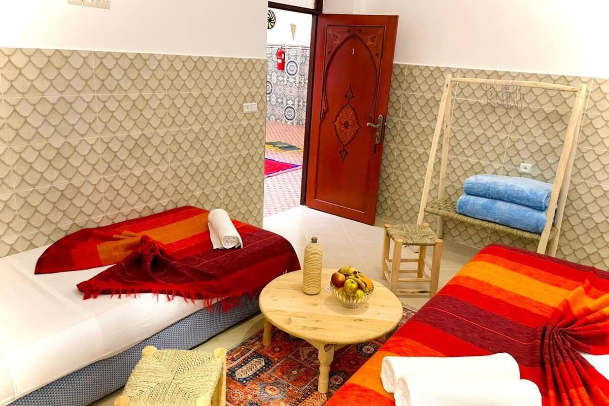 A room with two beds, a small table with a fruit bowl, a chair, and a hanging chair with towels.
