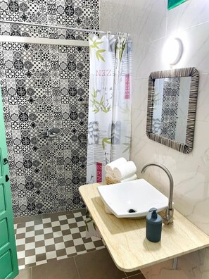 A bathroom with a checkered floor, a sink with a soap dispenser, a mirror with a patterned frame, and a shower area with patterned tiles and a bamboo shower curtain. - riad la joie (Marrakech)