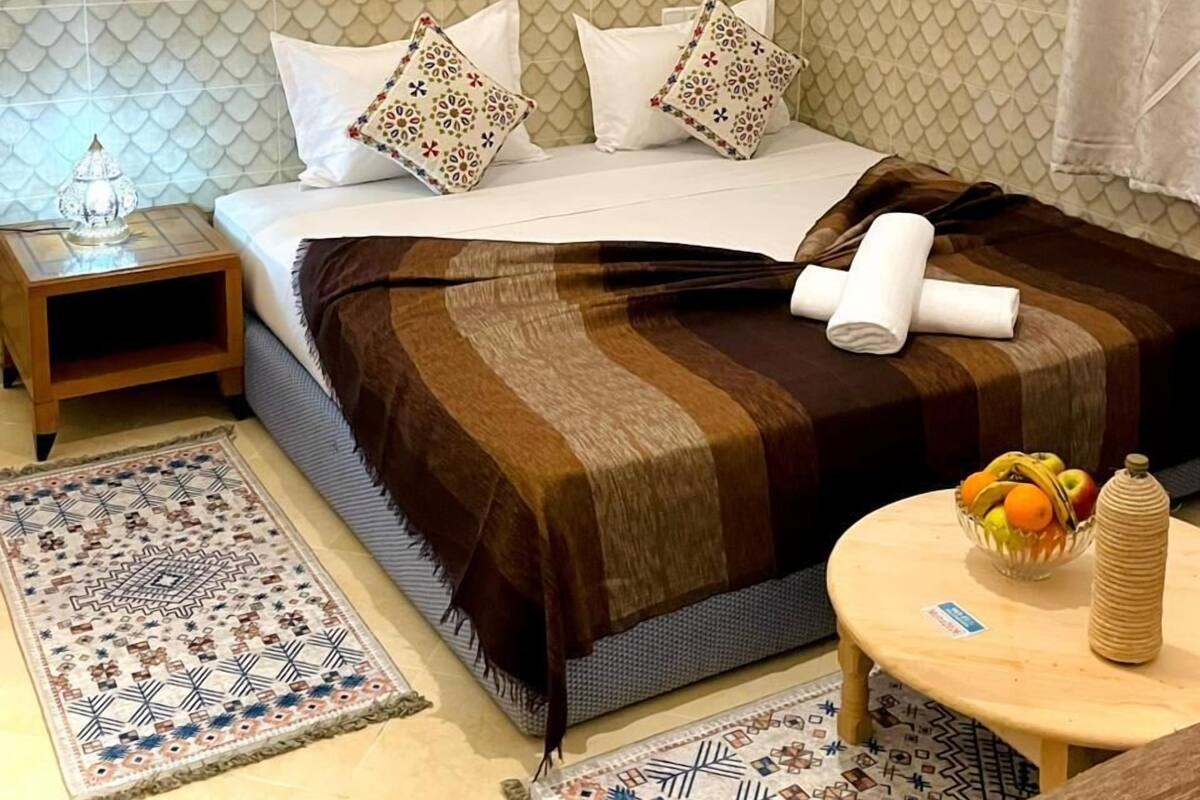 A hotel room with a bed, a nightstand, a small table with fruit, a woven basket, and a wall-mounted air conditioner.