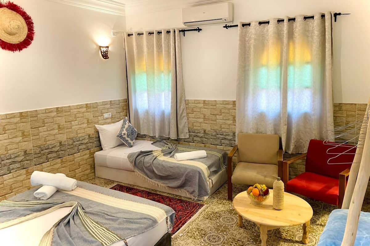 A hotel room with two beds, a sofa, a chair, a small table with fruit, and a wall-mounted air conditioner.