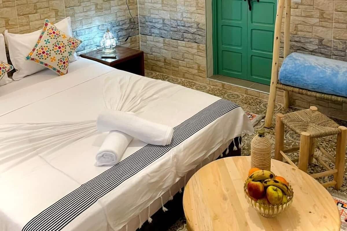 A hotel room with a bed, a round wooden table with fruit, a green door, and a wall-mounted light fixture.