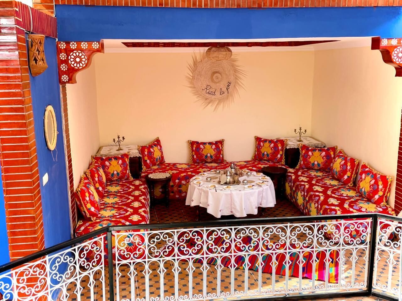 Un rincón amoblado con asiento, una mesa redonda, cojines estampados en rojo y blanco, y un tapiz decorativo.