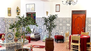 Una habitación con baldosas estampadas, una planta grande en maceta, un sofá rojo, una silla de madera y una mesa pequeña con mantel rojo.
