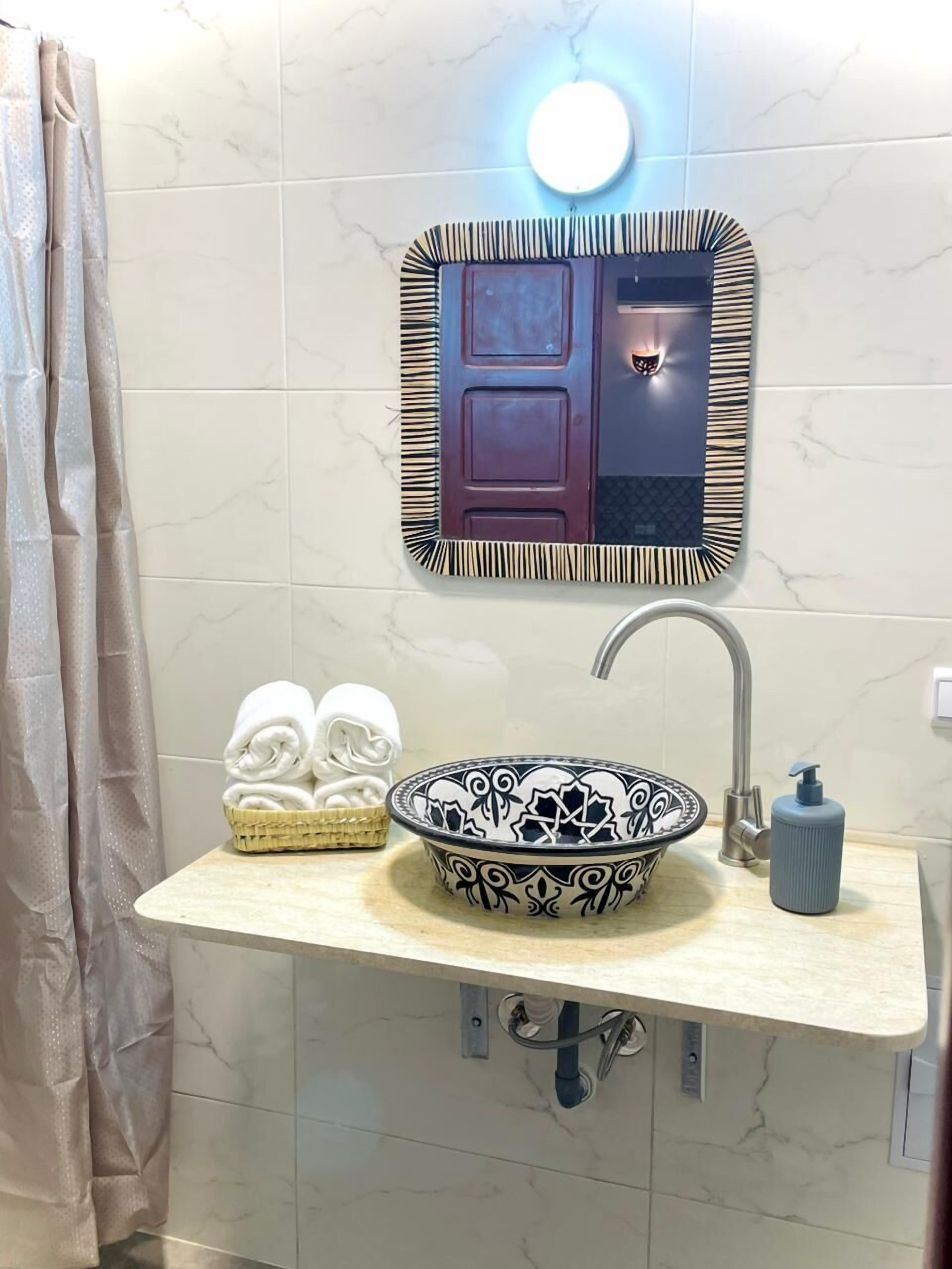 A bathroom with a marble-tiled wall, a sink with a decorative bowl, a mirror with a striped frame, a towel basket, and a shower curtain.