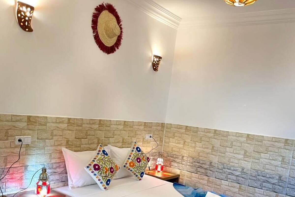 A bedroom with a brick-patterned headboard, a bed with blue bedding, decorative pillows, a wall-mounted hat, and a hanging light fixture.