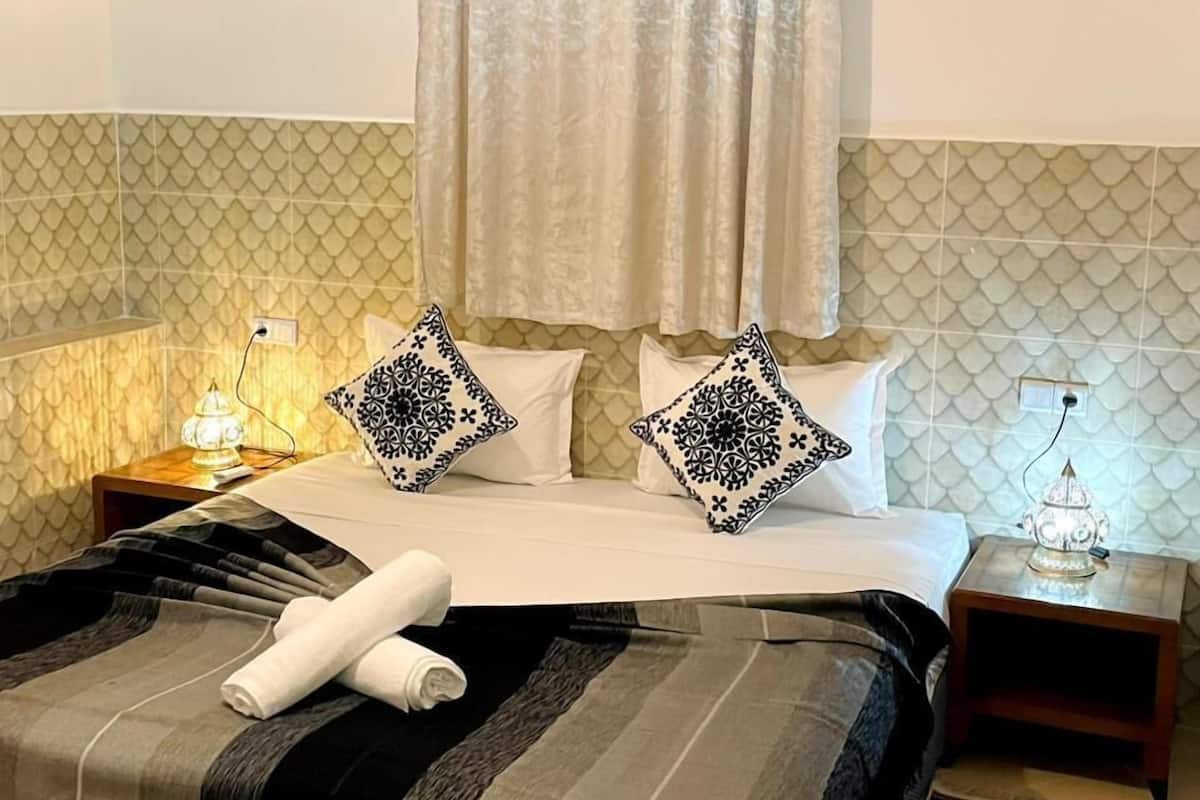 A hotel room with a bed, two bedside tables, a wall-mounted air conditioner, and a decorative wall piece.
