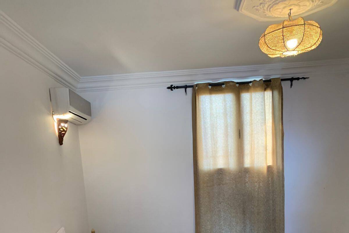 A double bed with a brown and white striped blanket, a small table with a checkered tablecloth, and a ceiling light fixture.