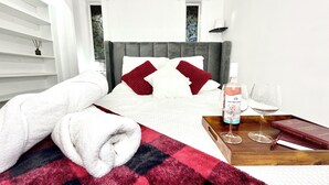 Een net opgemaakt bed met een rood-wit geruit dekbed, een fles rosé wijn, twee wijnglazen en een boek op een houten dienblad.