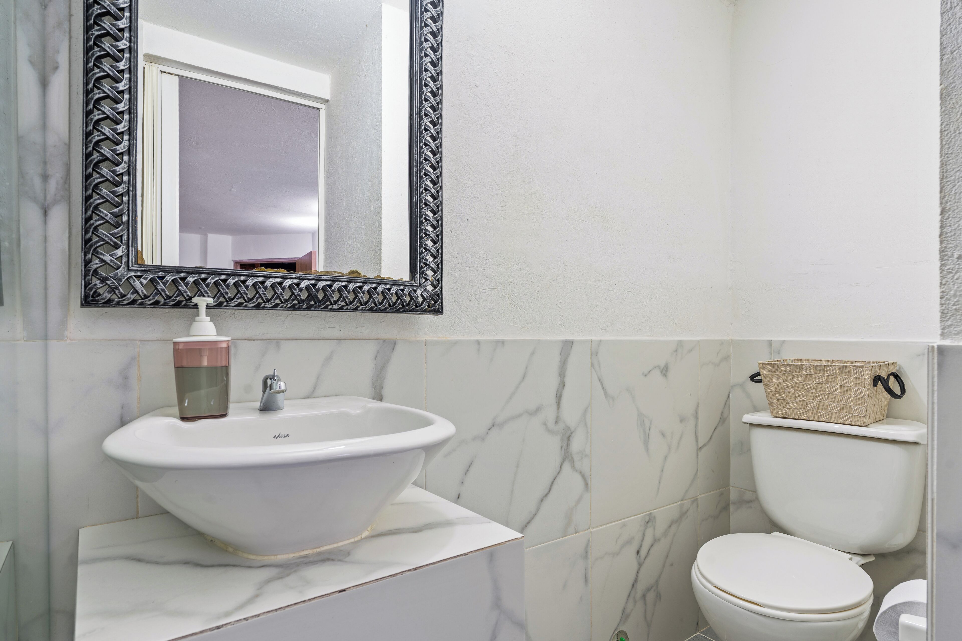Ein Badezimmer mit einem weißen Waschbecken, einem Spiegel mit schwarzem Rahmen und einer Toilette mit einem beigefarbenen Korb darauf.