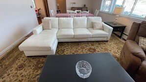 Ein Wohnzimmer mit einem weißen Ecksofa, einem Glastisch, einem braunen Sessel, einem Esstisch mit Stühlen und einem Fenster mit Jalousien.