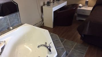 A hotel bathroom with a bathtub, a sink, a vanity with a mirror, and a bedside table with a lamp.