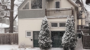 A two-story house with a balcony, a garage, and two snow-covered evergreen trees in the front yard. - Cozy Coach House - Step Outside to Frank Lloyd Wright Architectural Icons! (Oak Park)