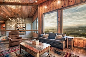 Ein großzügiger Wohnbereich mit großem Fenster und Blick auf die Berge, einer Ledersofa, einem Holztisch und einer hohen Decke mit freiliegenden Balken.