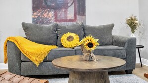 Un sofá gris con una manta amarilla, una mesa redonda de madera y un jarrón con girasoles.