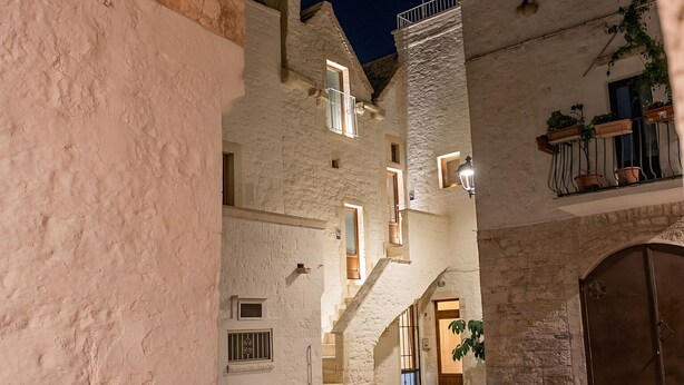 Une rue étroite bordée d’un bâtiment en pierre, doté d’un escalier, d’une porte en bois et d’un balcon orné de plantes en pot.