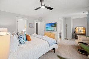 Une chambre à coucher comprenant un lit, un ventilateur de plafond, un téléviseur fixé au mur, une commode et une coiffeuse avec éclairage.