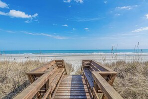 Ein hölzerner Steg führt zu einem Strand bei blauem Himmel.