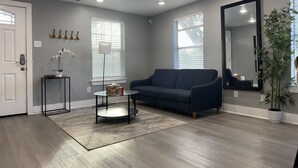A living room with a blue sofa, a round coffee table, and a large mirror. There are two windows with blinds, a potted plant, and a door with a coat rack. - Amazing 2-bedroom house in beautiful Houston with AC (Houston)