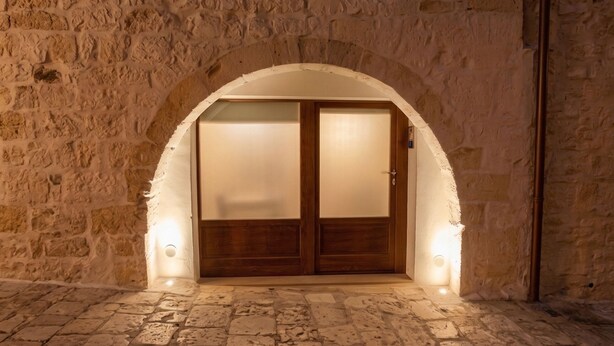 Un arco de entrada con una puerta de madera, iluminado por luces en la pared, que da paso a un interior con paredes de piedra.