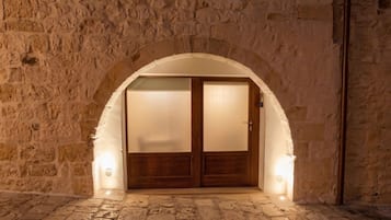 Un arco d'ingresso con una porta in legno, illuminato da luci a parete, che conduce a un interno con pareti in pietra.