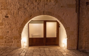 An arched doorway with a wooden door, illuminated by wall-mounted lights, leading into a stone-walled interior. - Arcobaleno - Campanile Inn; Comfortable room in historic building in Locorotondo (Locorotondo)