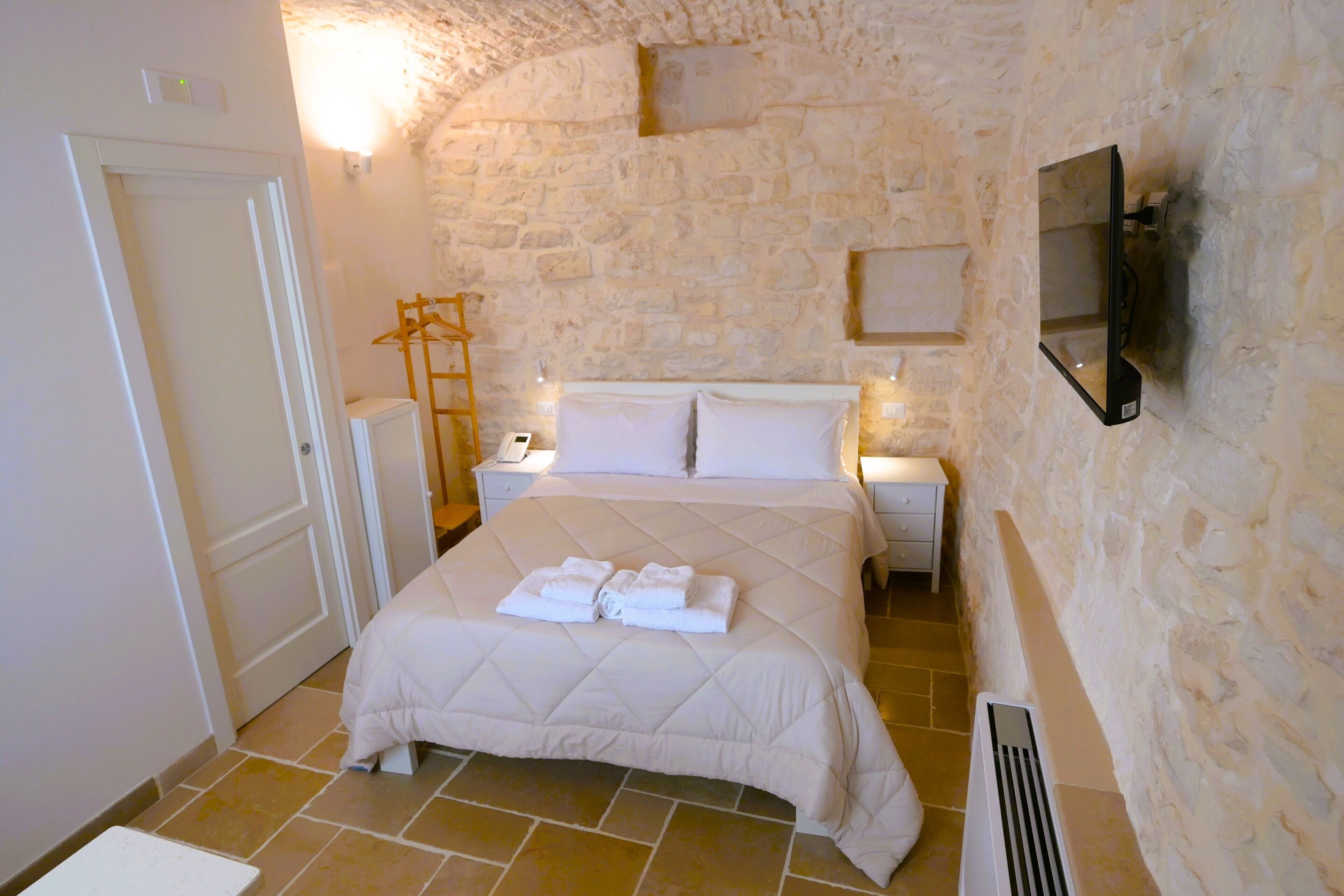 Una camera da letto con parete in pietra, un letto con biancheria bianca, una TV fissata al muro e un termosifone a pavimento.