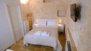 Un dormitorio con pared de piedra, una cama con ropa blanca, un televisor montado en la pared y un radiador en el suelo.