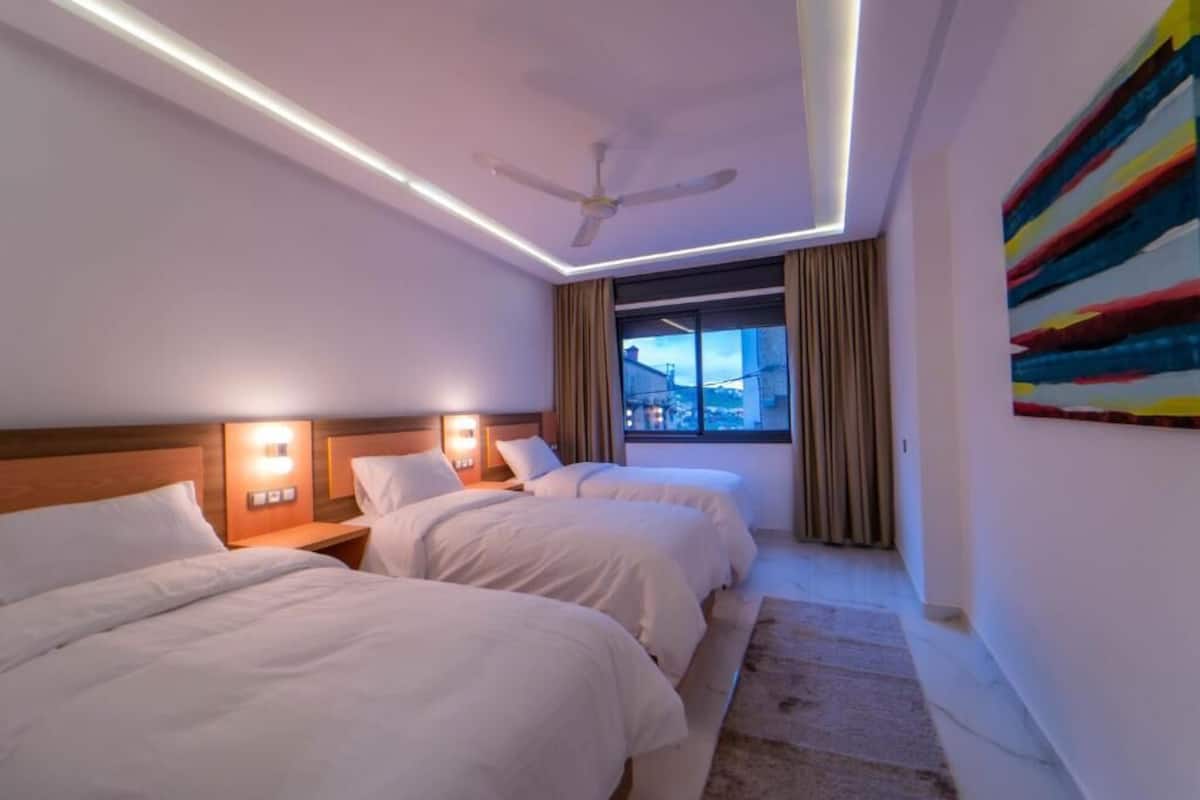 Habitación de hotel con dos camas, ventilador de techo, ventana grande con cortinas y un cuadro abstracto de colores en la pared.