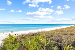 Ein Strand mit klarem, blauem Wasser, weißem Sand und vereinzelter Vegetation.