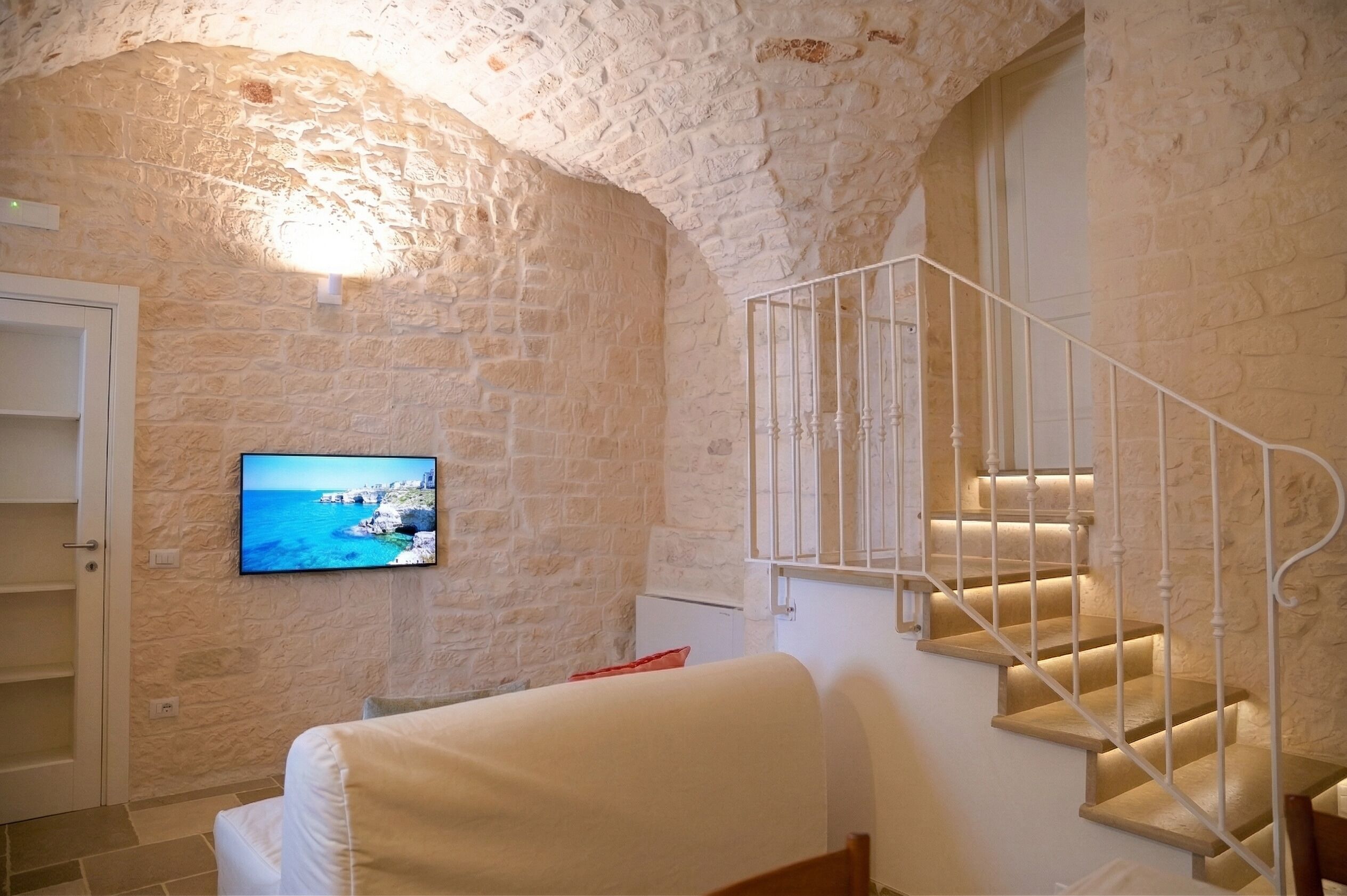 Una stanza con parete in pietra, televisore a muro, una scala con ringhiere bianche e un letto con biancheria da letto bianca.