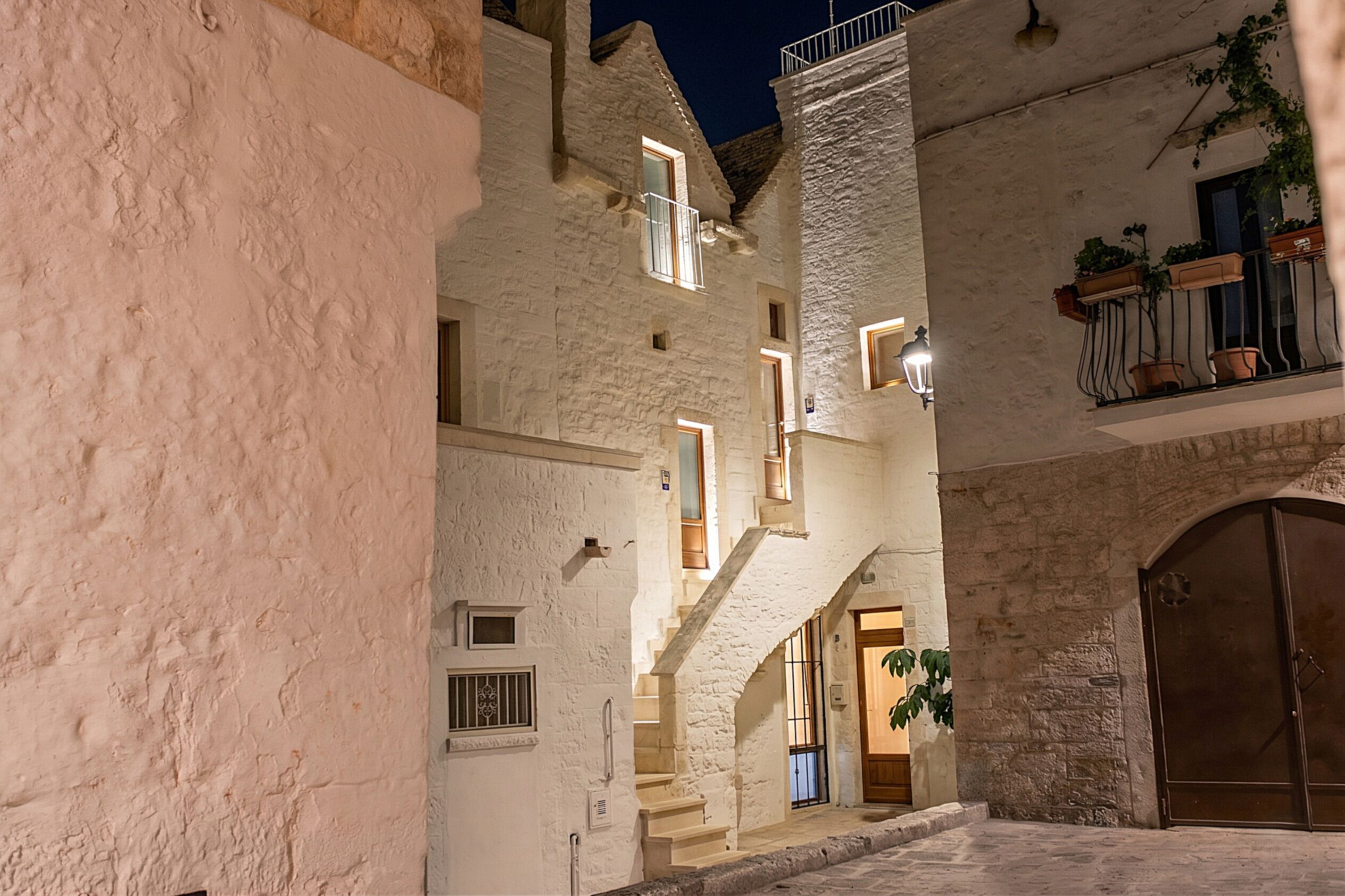 Un vicolo stretto con un edificio in pietra, caratterizzato da una scala, un portone in legno e un balcone con vasi di piante.