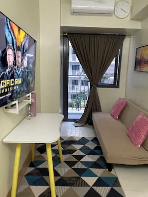 A living room with a sofa, a small table, a TV mounted on the wall, a window with curtains, and a patterned floor. - Cozy 1-bedroom condo in serene Pasay City with AC (Pasay City)