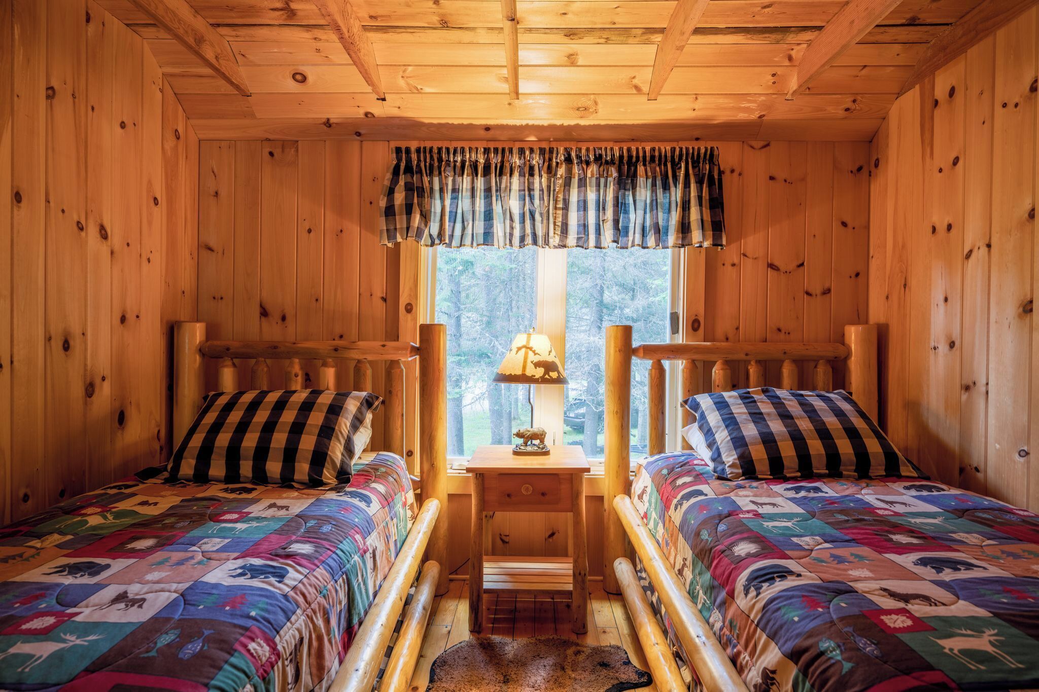 A cozy cabin room with two beds, a window with curtains, a lamp, and a small table.