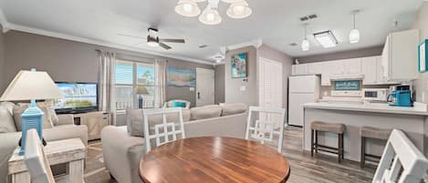 A modern living room with a round wooden table, a sofa, and a TV. There is a kitchen area with white cabinets and a bar.