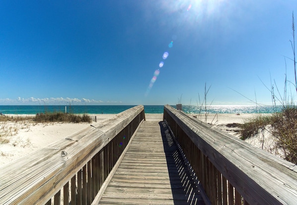 Ein hölzerner Steg führt zu einem Strand mit blauem Himmel und Blick auf das Meer.