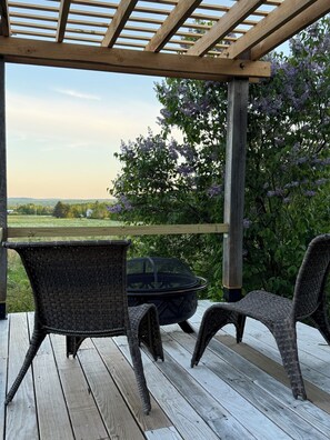 Una terraza de madera con sillas de mimbre y una mesa negra, con vistas a un jardín lleno de flores púrpuras.