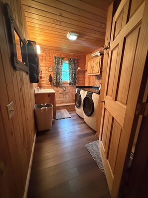 Een compact interieur met houtaccenten, voorzien van een wasmachine, een klein wastafeltje en een raam met gordijnen.