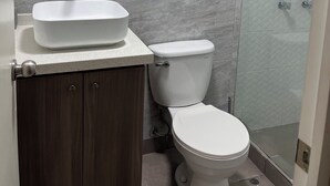 Ein Badezimmer mit einem weißen Waschbecken, einer Toilette und einem Duschbereich. Über der Toilette befinden sich Holzregale.