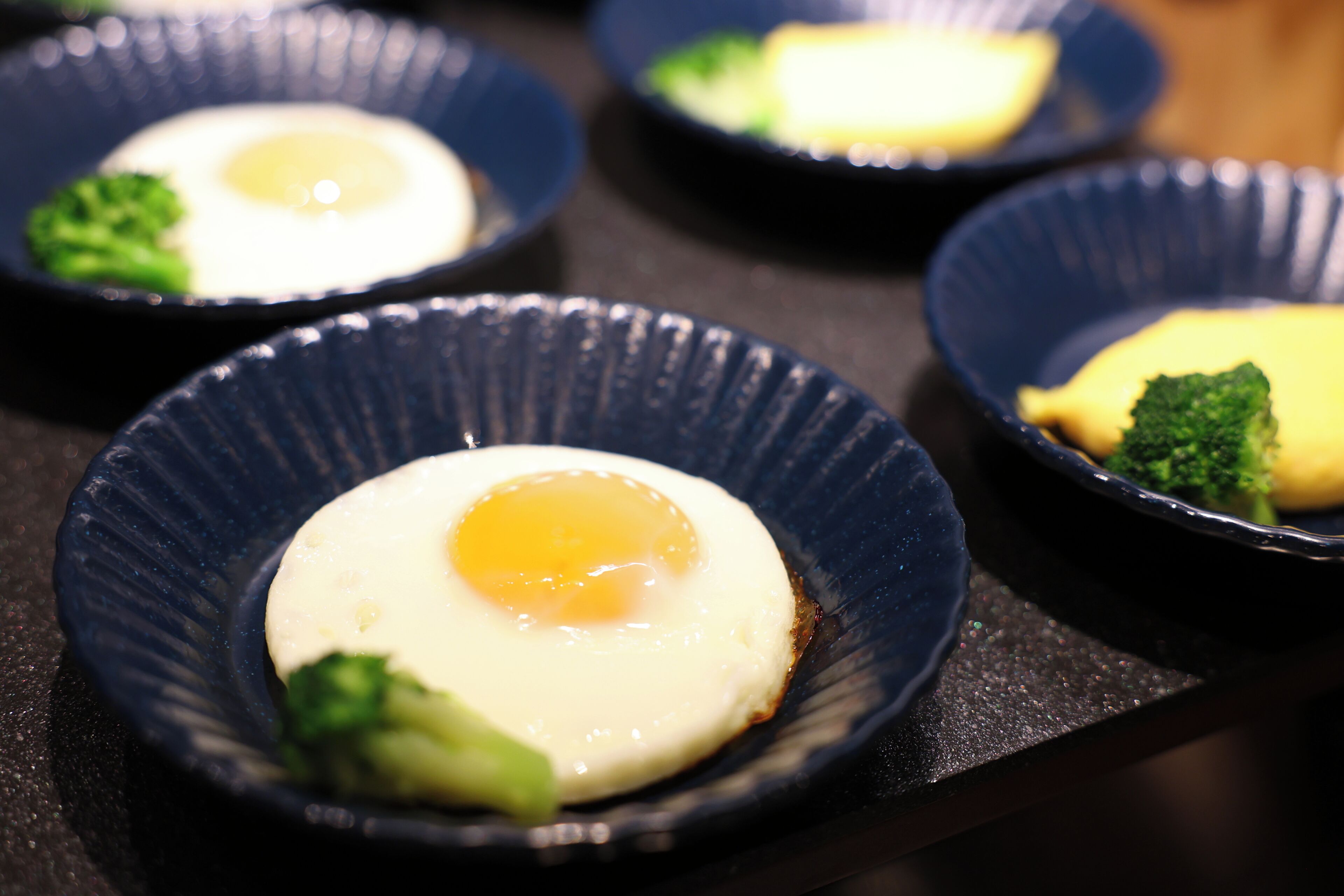 A series of dark blue bowls containing a fried egg and broccoli.